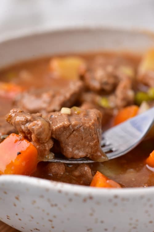 A closeup of a fork holding a piece of beef over a bowl of vegetable beef soup.