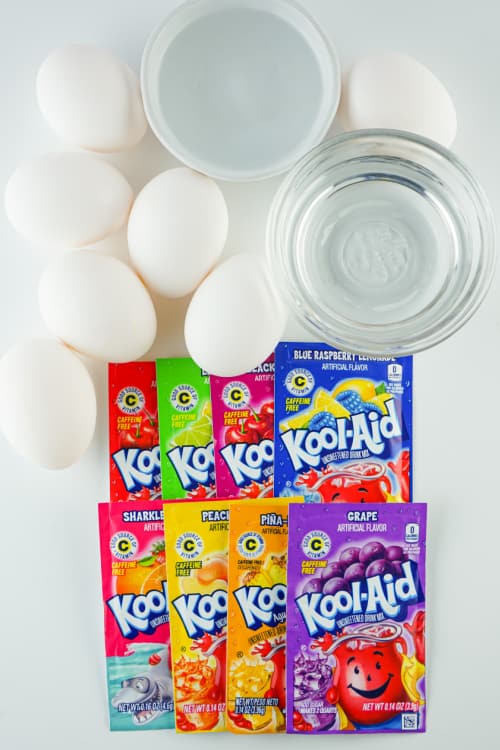 An overhead view of several packets of Kool-Aid powder alongside several eggs and a small glass bowl of water.