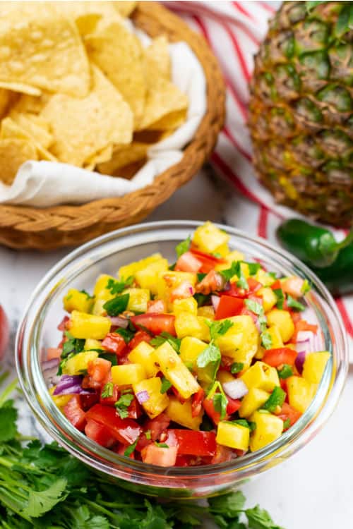 A small glass bowl of pineapple pico de gallo alongside a pineapple and bowl of tortilla chips on a marble countertop.
