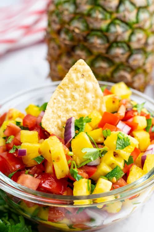 A tortilla chip being dipped into a small glass bowl of pineapple pico de gallo.