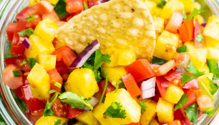 A closeup of a tortilla chip being dipped into a small glass bowl of pineapple pico de gallo.