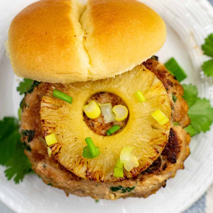 An overhead view of a chicken teriyaki burger topped with a slice of pineapple and garnished with cilantro and green onions on a white plate.