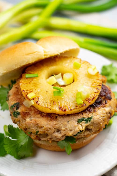 A chicken teriyaki burger topped with a slice of pineapple and garnished with cilantro and green onions on a white plate.