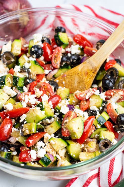 A wooden spoon in a large glass bowl of Mediterranean cucumber salad.