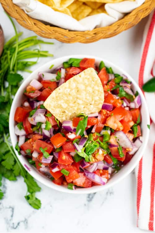 An overhead view of a tortilla chip being dipped into a white bowl of pico de gallo.