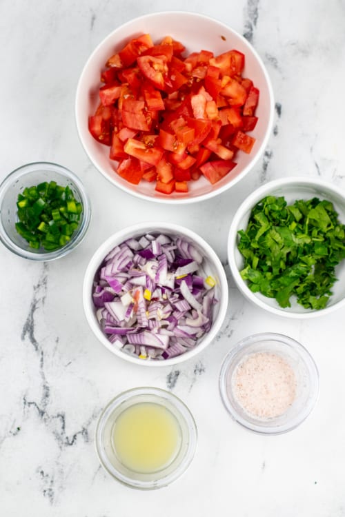 An overhead view of several ingredients for pico de gallo on a marble countertop.