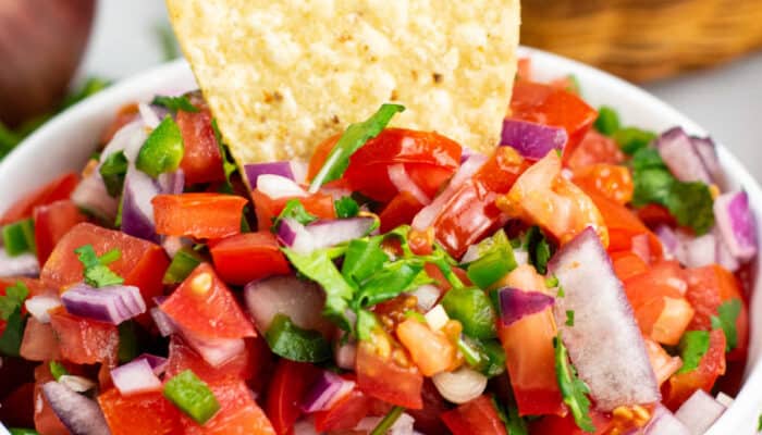 A close-up of a tortilla chip being dipped into a white bowl of pico de gallo.