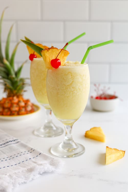 Two piña colada mocktails topped with pineapple and cherry on a marble countertop.