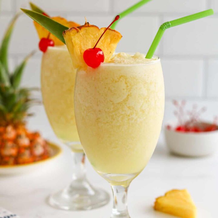 Two piña colada mocktails topped with pineapple and cherry on a marble countertop.