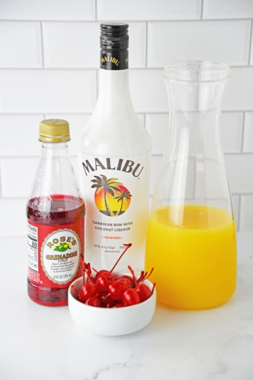 Several ingredients for malibu rum punch in front of a white brick background.