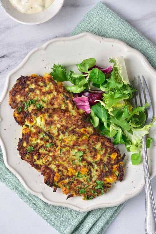 An overhead view of a plate of zucchini corn fritters with a small side salad.