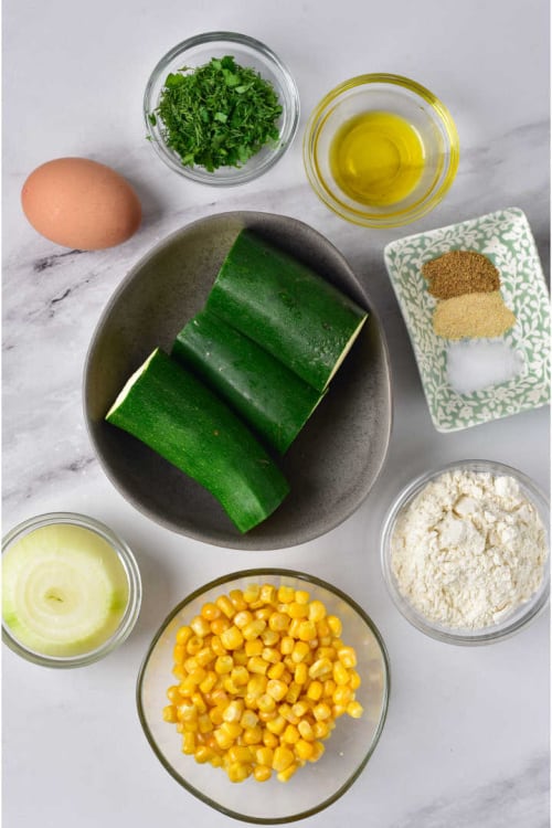 An overhead view of several ingredients for zucchini corn fritters on a marble countertop.