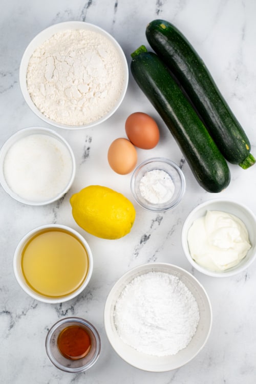 The ingredients for lemon zucchini bread on a white marble background. Zucchini, flour, sugar , eggs, lemon juice, lemon zest, baking powder, greek yogurt, oil, powdered sugar and vanilla extract