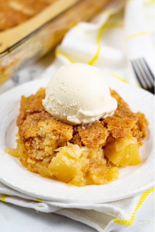 A small white plate of pineapple dump cake topped with a scoop of vanilla ice cream.