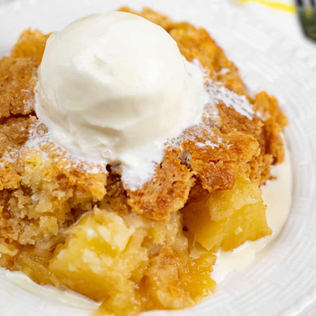 A close-up of a small white plate of pineapple dump cake topped with a scoop of vanilla ice cream.