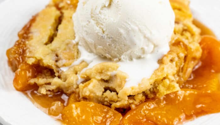 A close up of a piece of peach cobbler on a white plate with a scoop of vanilla ice cream on top