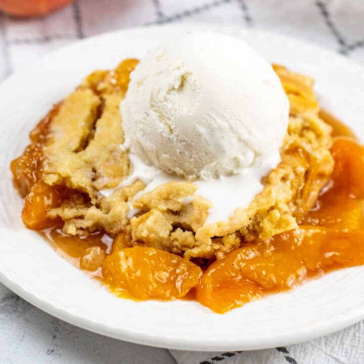 A close up of a piece of peach cobbler on a white plate with a scoop of vanilla ice cream on top