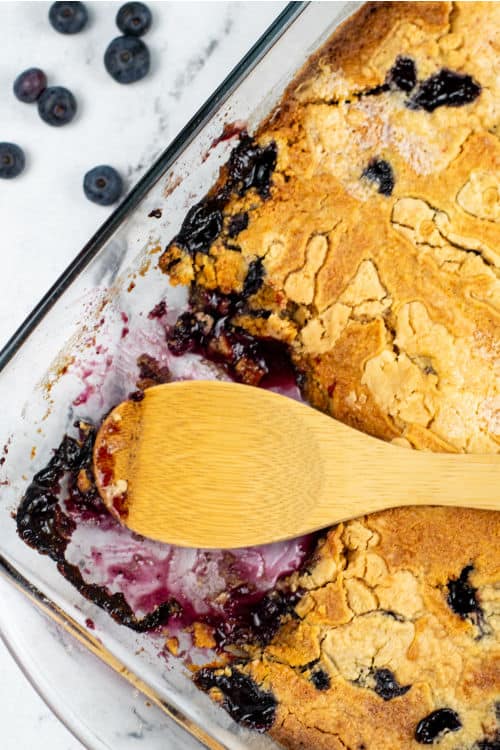A clear glass baking dish with blueberry cake mix cobbler in it, a portion has been taken out with a wooden spoon.
