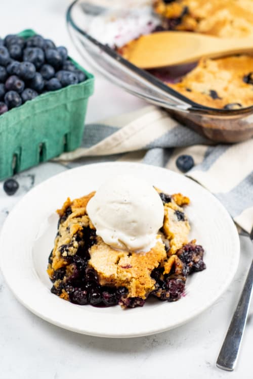 A white plate with a serving of blueberry cobbler on it. The cobbler has been topped with a scoop of vanilla ice cream.