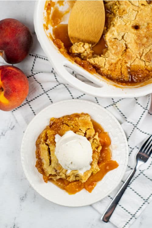 A piece of peach cobbler with cake mix on a white plate topped with a scoop of vanilla ice cream