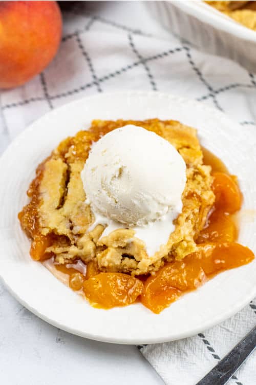 A white plate with a piece of peach cobbler topped with a scoop of vanilla ice cream