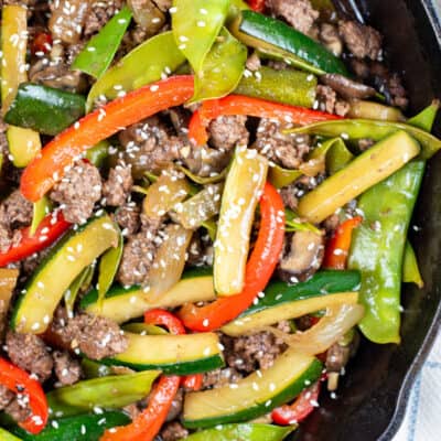 A close up picture of a cast iron skillet full of ground beef stir fry. It is ground beef peppers, onions, zucchini and snow peas