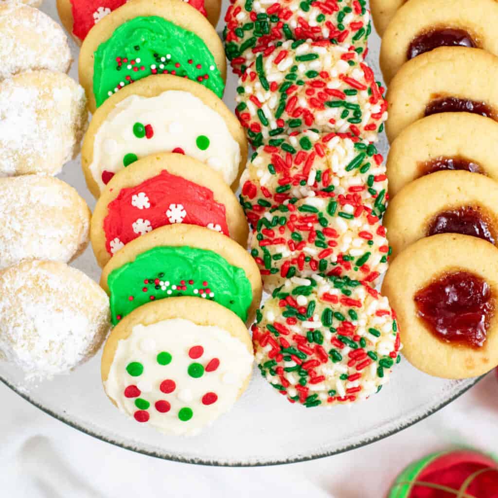 A close up of a plate of cookies. Thumbprint cookies, sprinkle cookies, frosted sugar cookies and powdered sugar coated cookies